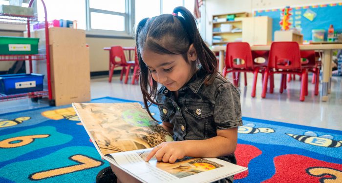 A young child in classroom sitting on a rug looking at a picture book.
