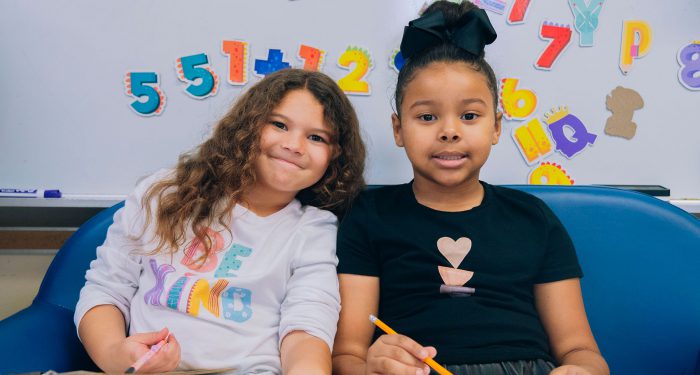 Two young children sit on a small couch in a classroom.