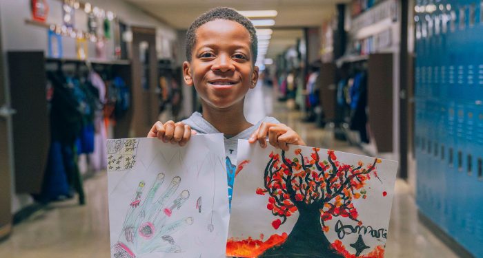 A student in a school hallway smiles while holding up their artwork.